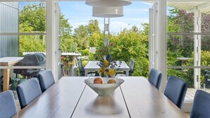 A dining area with a large table and chairs, a hanging light fixture, and a view of a garden through large windows.