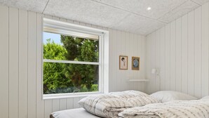A bedroom with a bed, a window with a view of greenery, and a wall-mounted shelf with two framed pictures.
