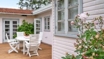 En uteplats med ett bord och stolar, en vit byggnad med fönster och en rosenbuske i förgrunden.