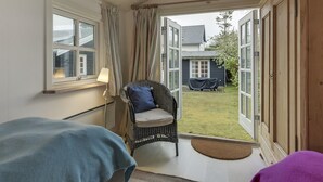 Une pièce avec un fauteuil en osier, une petite table et une vue sur un jardin à travers des portes ouvertes.