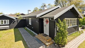 Une petite cabane de couleur sombre, dotée d’une cheminée, d’un petit porche et d’une porte ornée d’une fenêtre en forme de losange.