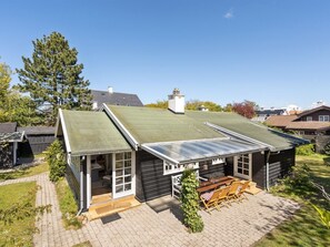 Ett enplanshus med ett grönt tak, ett glashus och en uteplats med utemöbler.