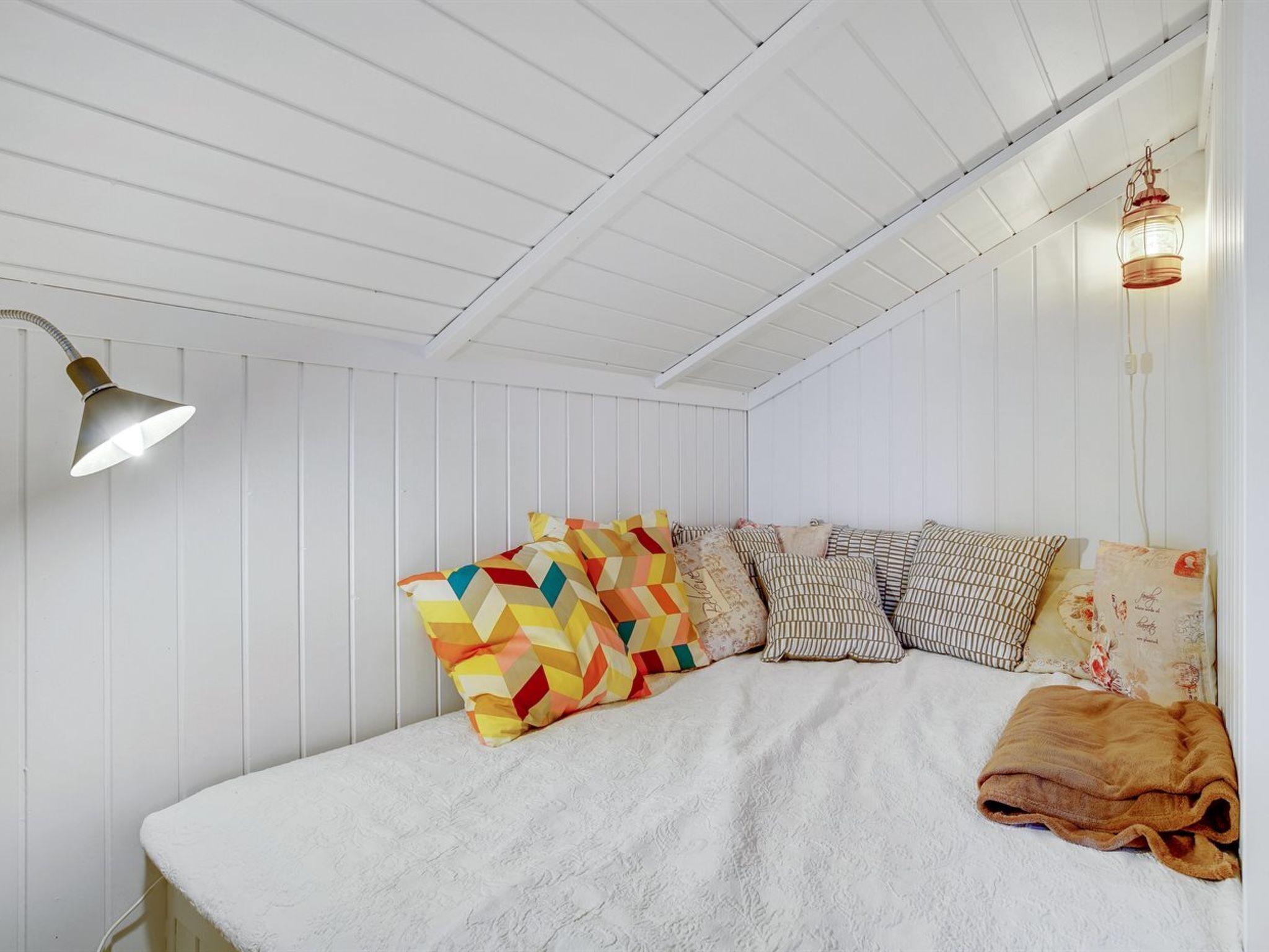 Et soveværelse med en hvid seng, farverige puder, et hvidt tæppe, en lampe og en hængende lysarmatur.