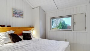 Un dormitorio con cabecera de madera, cama con ropa de cama blanca, ventana con vista a árboles y pared blanca con un cuadro colorido.