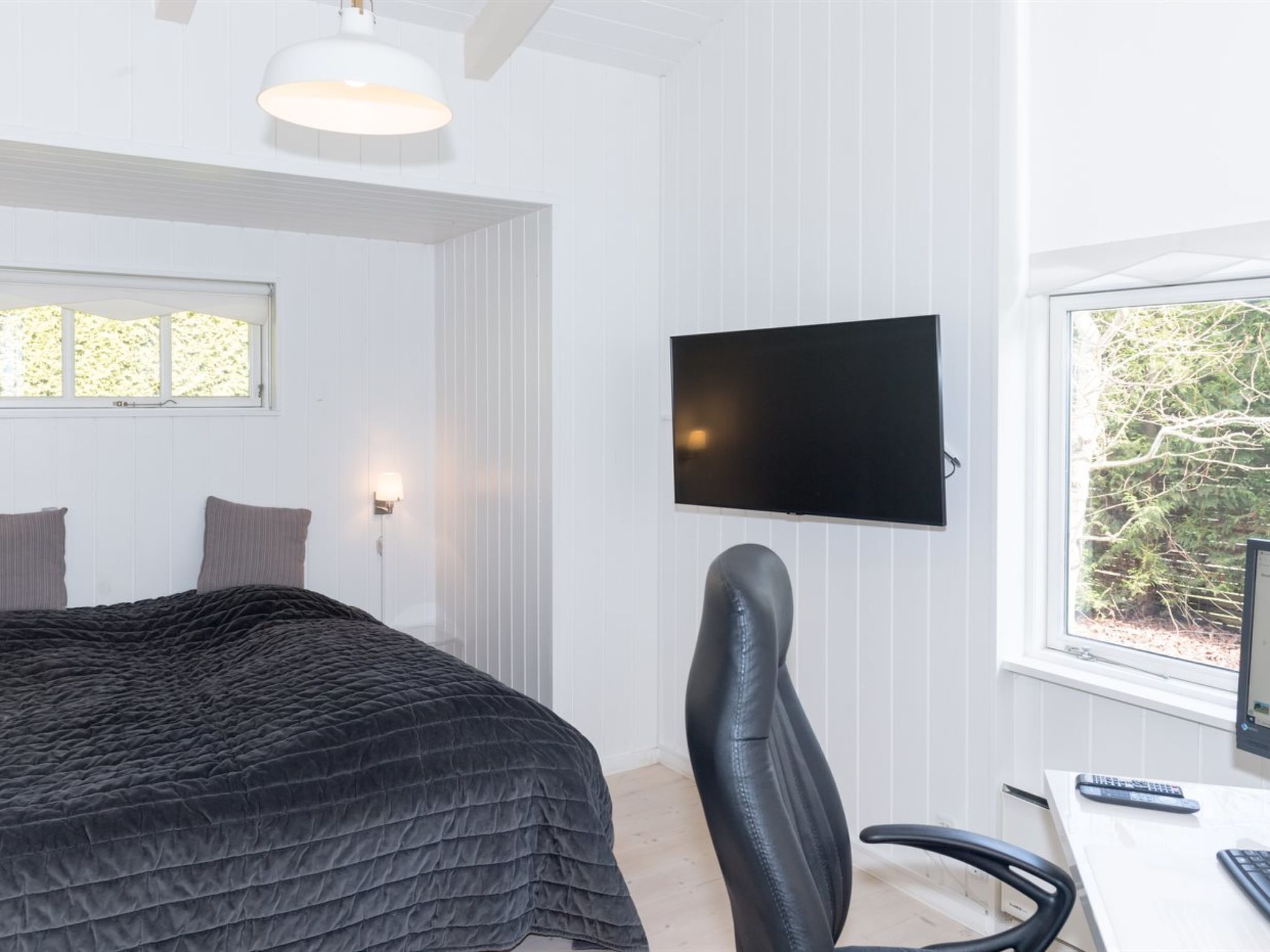 A bedroom with a bed, a desk with a computer, a chair, a TV, and a window with a view of trees.