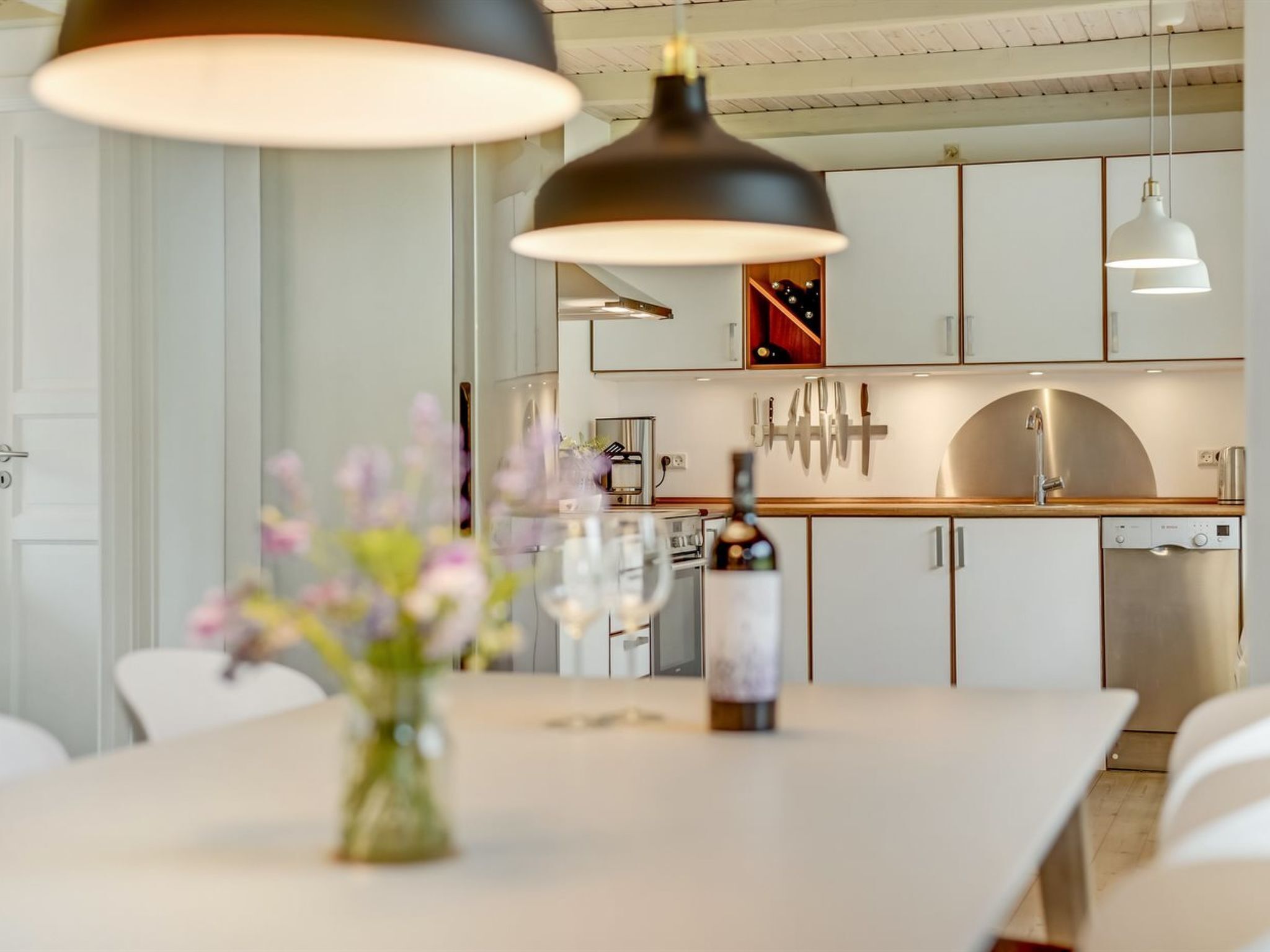 Une cuisine moderne avec des armoires blanches, un comptoir en bois et des appareils électroménagers en acier inoxydable. On aperçoit une table à manger avec un vase de fleurs au premier plan.