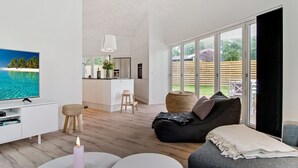 A modern living room with a TV, a white sofa, a black sofa, and a dining area with a kitchen in the background.