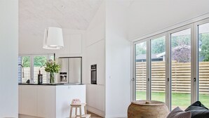 A modern kitchen with a central island, a dining area with a dark sofa and woven ottoman, and a large sliding glass door leading to a backyard.