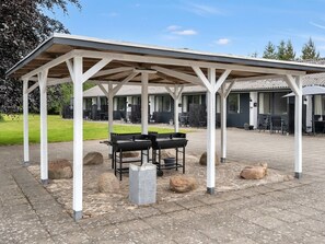 Ein überdachter Außenbereich mit Grill, Steinsitzgelegenheiten und Blick auf ein Gebäude mit zahlreichen Fenstern und Türen.