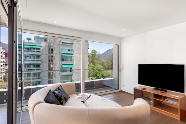 A modern living room with a large TV, a sofa, and a glass door leading to a balcony with a city view.