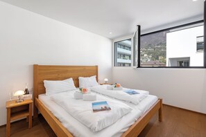 A hotel room with a wooden bed, white linens, a nightstand with a bottle and glasses, and a view of buildings through a large window. - Appartamento Lago e Monti - Garage & Terrace in Locarno (Locarno)