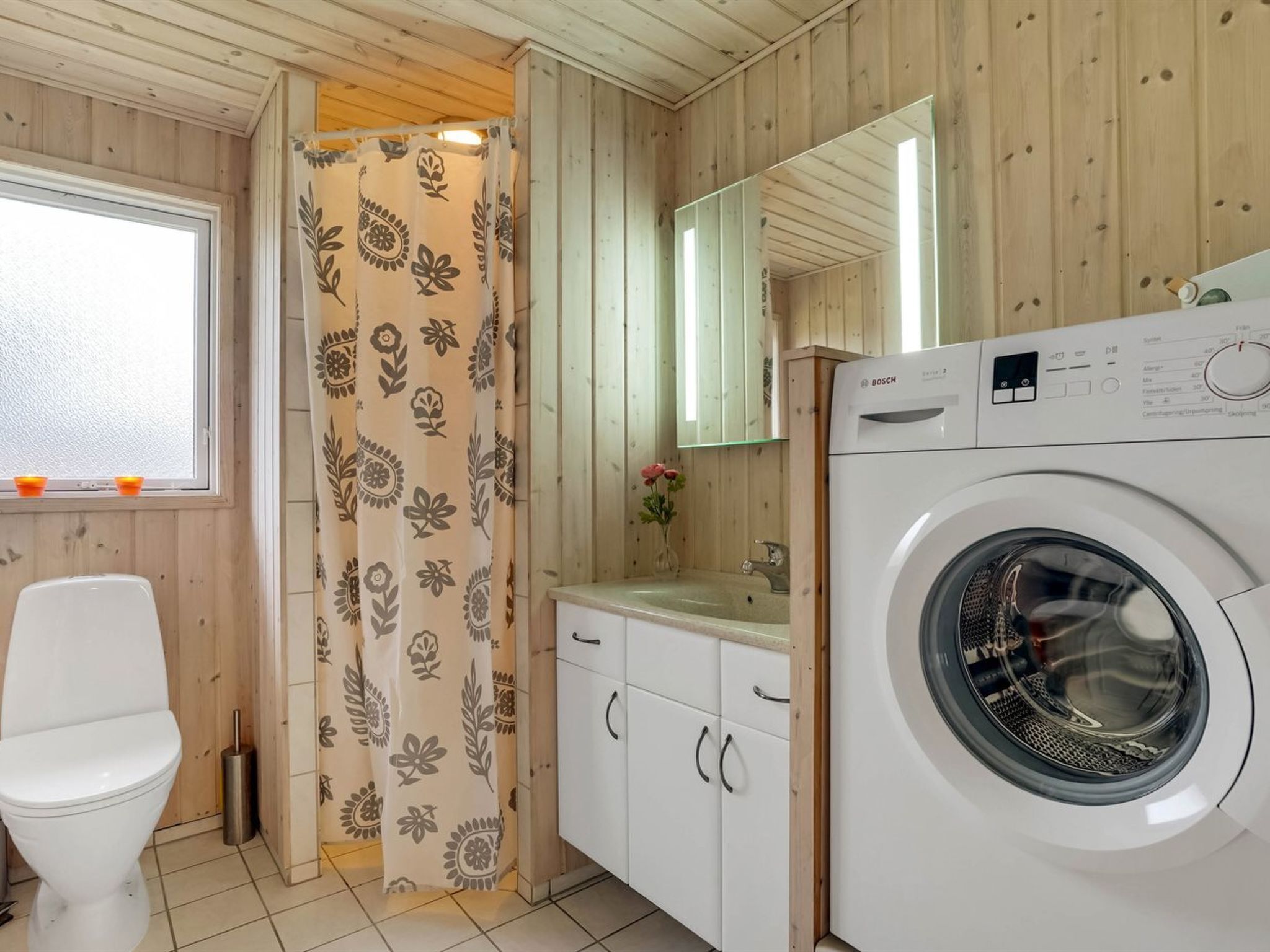 A compact, wooden-finished bathroom with a washing machine, a toilet, and a shower curtain with a floral pattern.