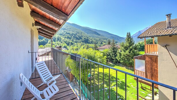 Ein Balkon mit Holzgeländer, einem weißen Stuhl und Blick auf eine grüne Landschaft mit Bergen.