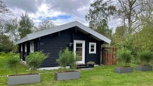 A small, dark-colored house with a white door and windows, surrounded by a grassy yard and several potted plants. - "Freyja" - 800m from the sea by Interhome (Væggerløse)