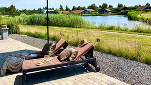 Un fauteuil de relaxation doté d’un auvent, une table avec une bouteille et des verres, et un chemin de gravier menant à un plan d’eau, avec des maisons en arrière-plan.