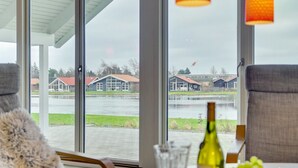 Une pièce dotée d’une grande fenêtre donnant sur un plan d’eau et des bâtiments. On y trouve une table avec une bouteille, des verres et des fruits.