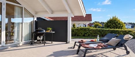 A covered outdoor area with a grill, lounge chairs, and a table with food and drinks.