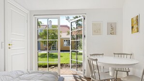 Un dormitorio con cama, una mesa de comedor con sillas, una puerta corrediza de vidrio que da a un jardín y una pared blanca con cuadros enmarcados.