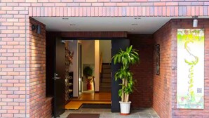 A brick exterior with a potted plant in the entrance, a door leading inside, and a sign with Japanese characters. - Guest House Siesta (Tokyo)