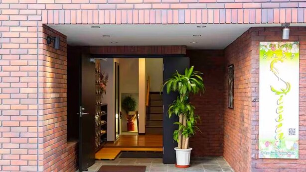 A brick exterior with a potted plant in the entrance, a door leading inside, and a sign with Japanese characters. - Guest House Siesta (Tokyo)