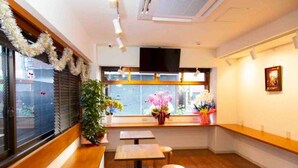 A restaurant interior with wooden tables and benches, a large window with blinds, and a wall-mounted TV. - Guest House Siesta (Tokyo)