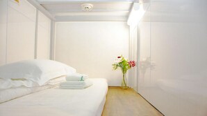 A neatly made bed with white linens and a folded towel, a small vase with flowers, and a wooden floor. - Guest House Siesta (Tokyo)