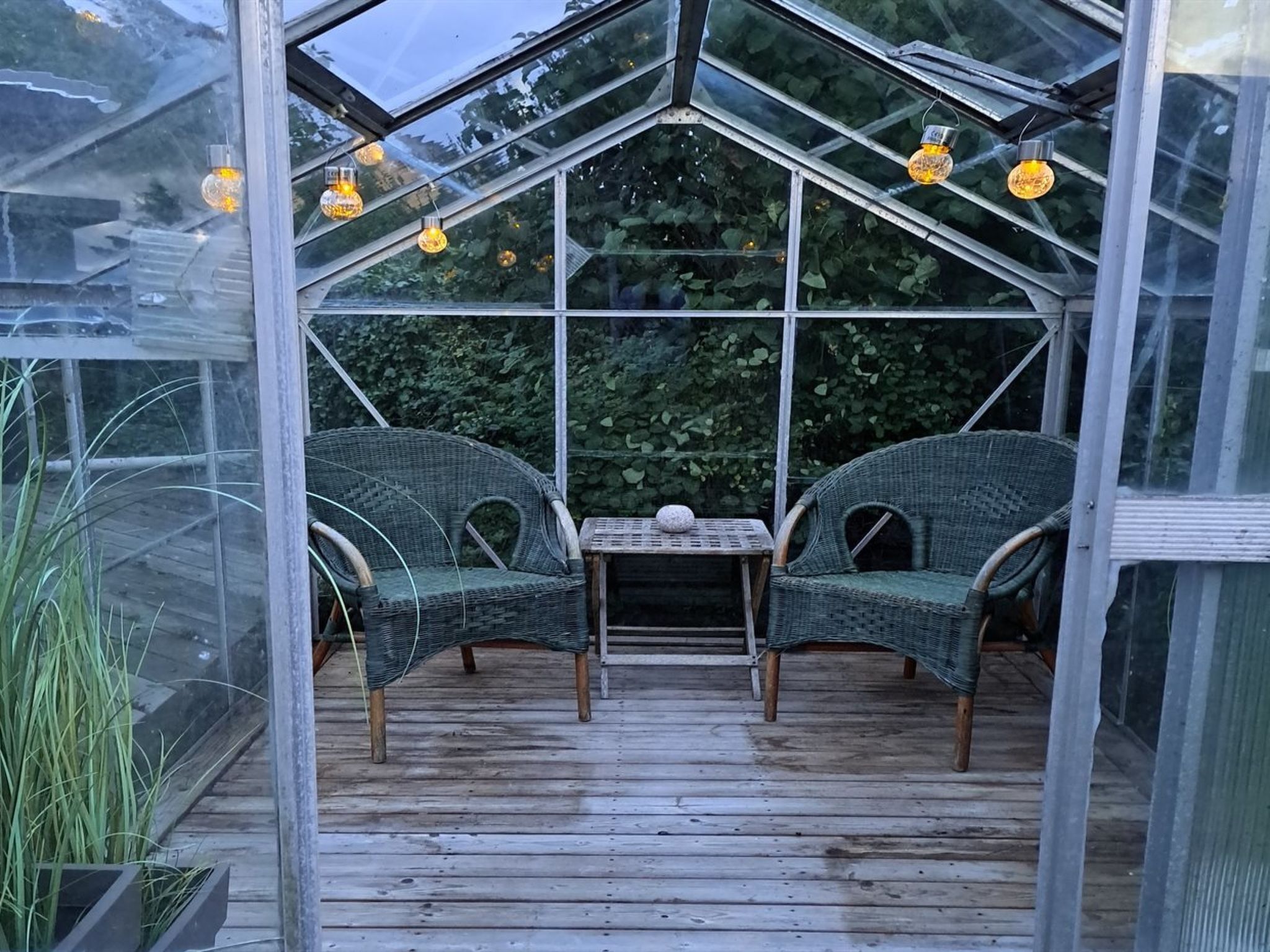 A greenhouse with two wicker chairs and a small table, surrounded by glass panels and wooden flooring.