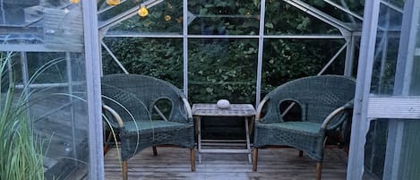 A greenhouse with two wicker chairs and a small table, surrounded by glass panels and wooden flooring.
