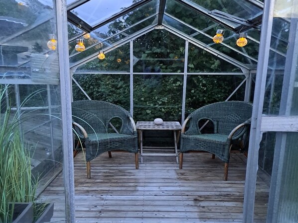 A greenhouse with two wicker chairs and a small table, surrounded by glass panels and wooden flooring.