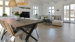 A spacious living room with a large wooden dining table, a sofa set, and a television.