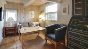 Una habitación acogedora con paneles de madera, techo de madera, un sofá, un sillón a rayas, una mesa con lámpara y un aparador con cajones.
