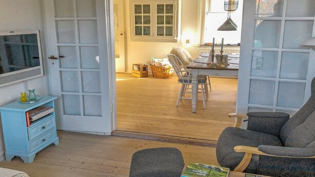 Een kamer met een televisie, een blauwe ladekast, een grijze bank en een eethoek met een tafel en stoelen.