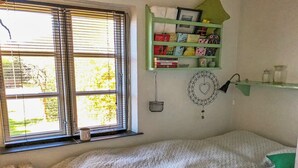 Een gezellige slaapkamer met een groot raam, een groene boekenkast, een aan de muur bevestigde dreamcatcher en een radiator.