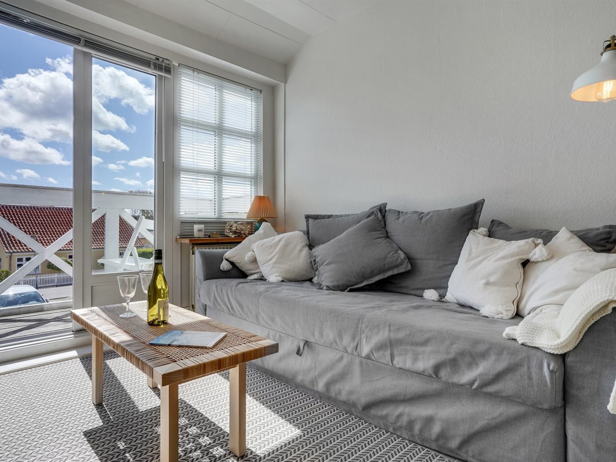 En moderne stue med en grå sofa, et træbord og udsigt til et hvidt hegn og et rødt tegltag gennem store vinduer.