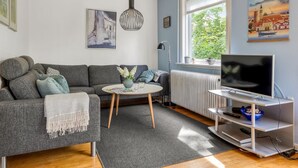 Een moderne woonkamer met een grijze bank, een ronde salontafel, een tv op een witte kast, een radiator en een raam met uitzicht op groen.