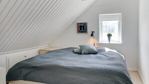 Ein Schlafzimmer mit schrägem Dach, einem Bett mit gesteppter Decke, einer Nachttischlampe, einem Fenster mit Ausblick und einem gerahmten Bild an der Wand.