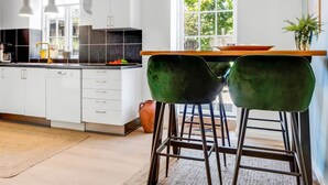 A modern kitchen with white cabinets, a black backsplash, and a window with a view of greenery. There are two green bar stools and pendant lights above the counter. - "Viivi" - 900m from the sea by Interhome (Skagen)
