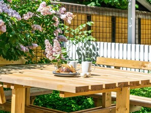 A wooden picnic table with a plate of pastries and two white cups, set against a backdrop of blooming lilac bushes and a white fence.