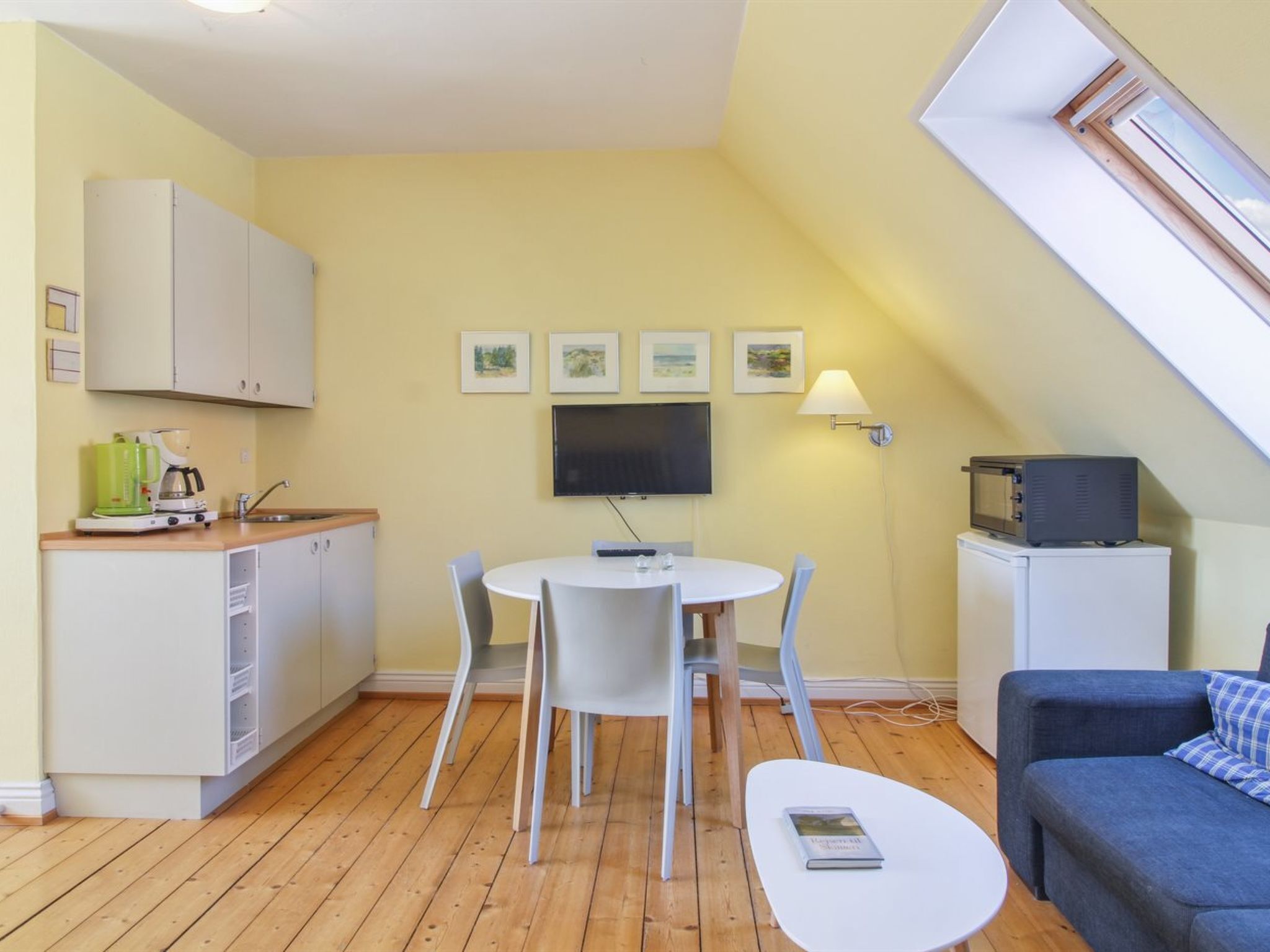 Un espace de vie compact comprenant un coin cuisine, un coin repas avec une table ronde et des chaises, un canapé et un téléviseur fixé au mur.