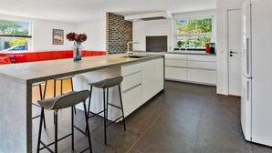 A modern kitchen with a central island, bar stools, a built-in oven, and a large window offering a view of greenery.
