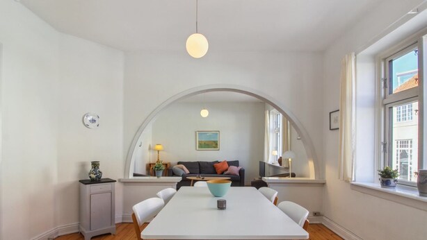 A dining area with a white table and chairs, a hanging light fixture, and a view through an arched window to a living room with a sofa and a painting. - "Gyrth" - 250m from the sea by Interhome (Skagen)