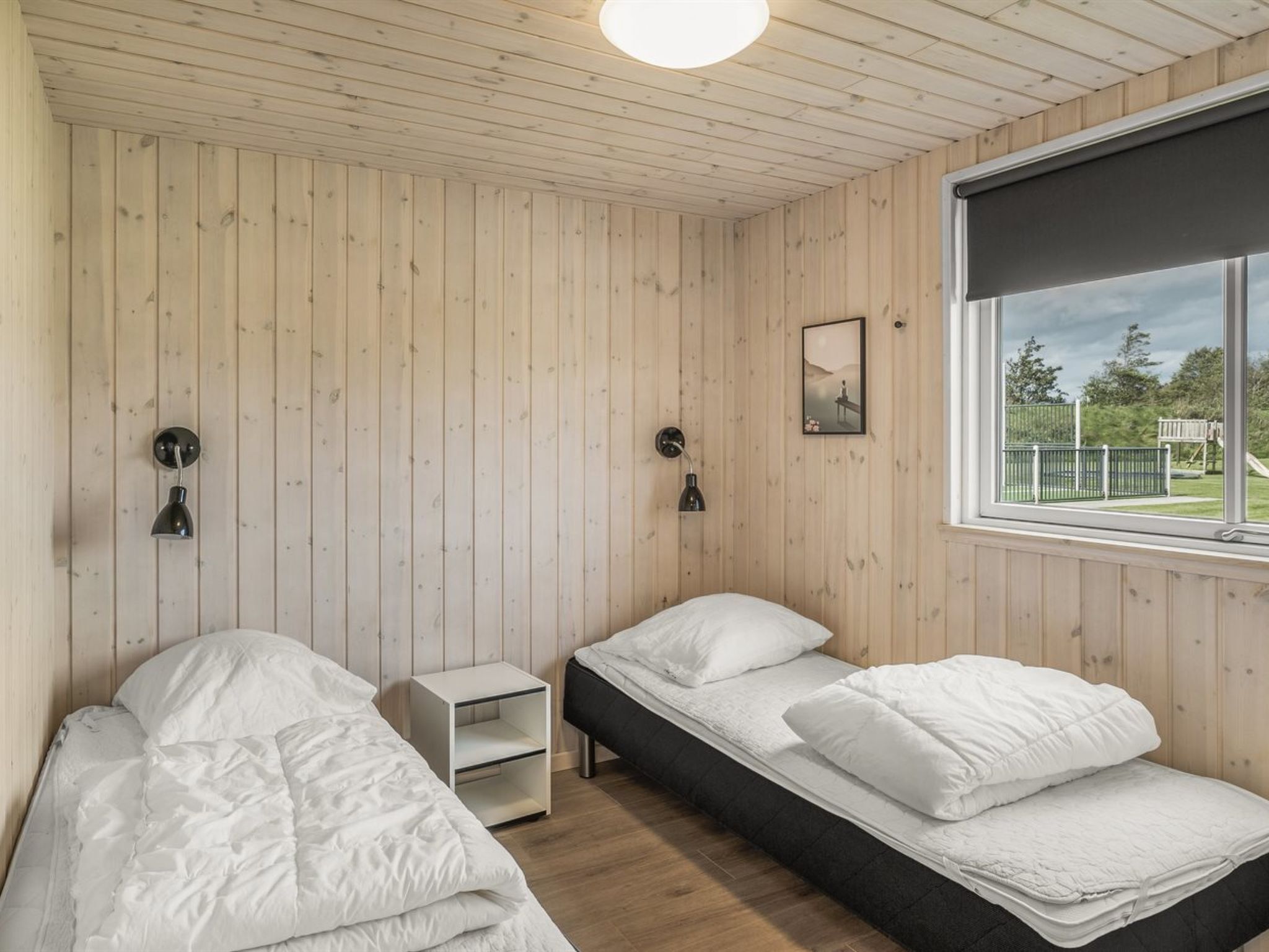 A room with two beds, a small white nightstand, a wall-mounted lamp, and a window with a view of a playground.
