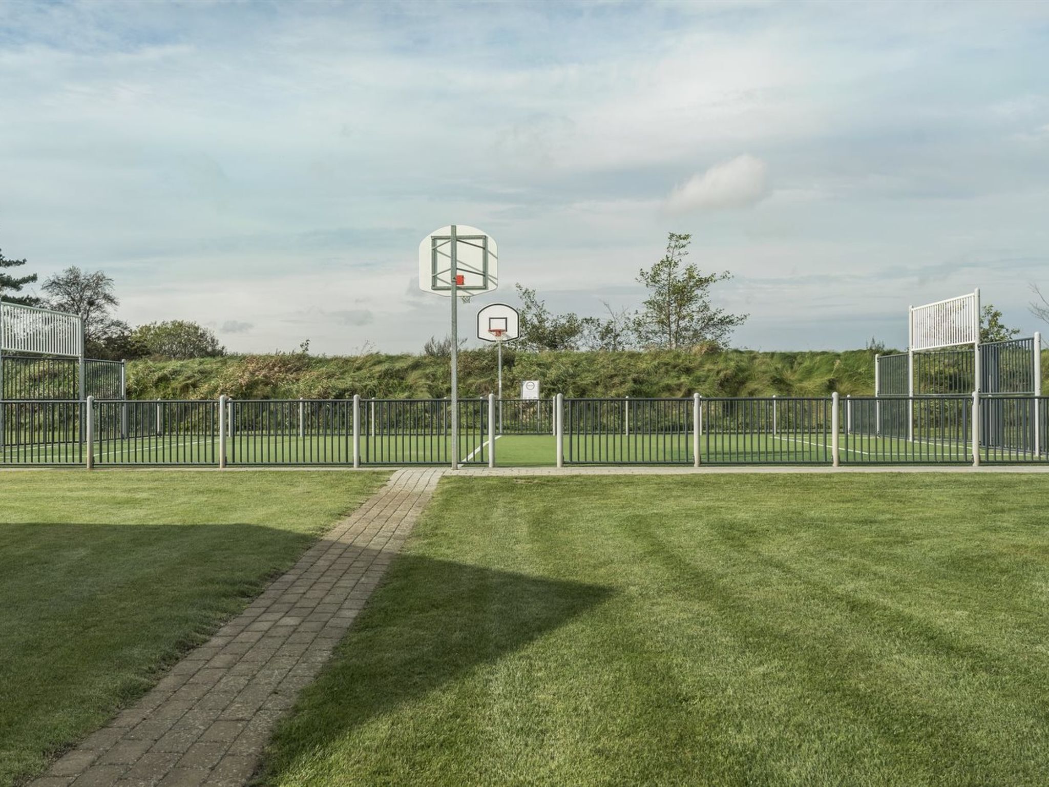 Una cancha deportiva con un aro de baloncesto, rodeada por una valla y un sendero pavimentado.