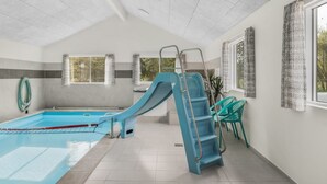 Una piscina interior con un tobogán azul, paredes de azulejos y ventanas con cortinas.