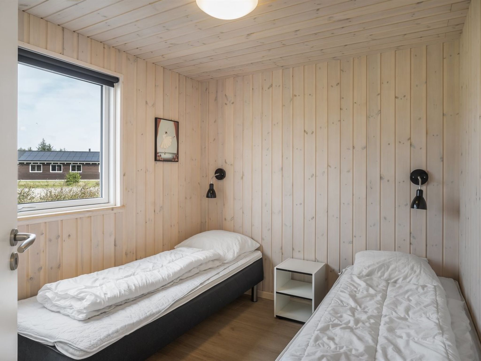 A bedroom with two single beds, a window, a small bedside table, and a wall-mounted lamp.