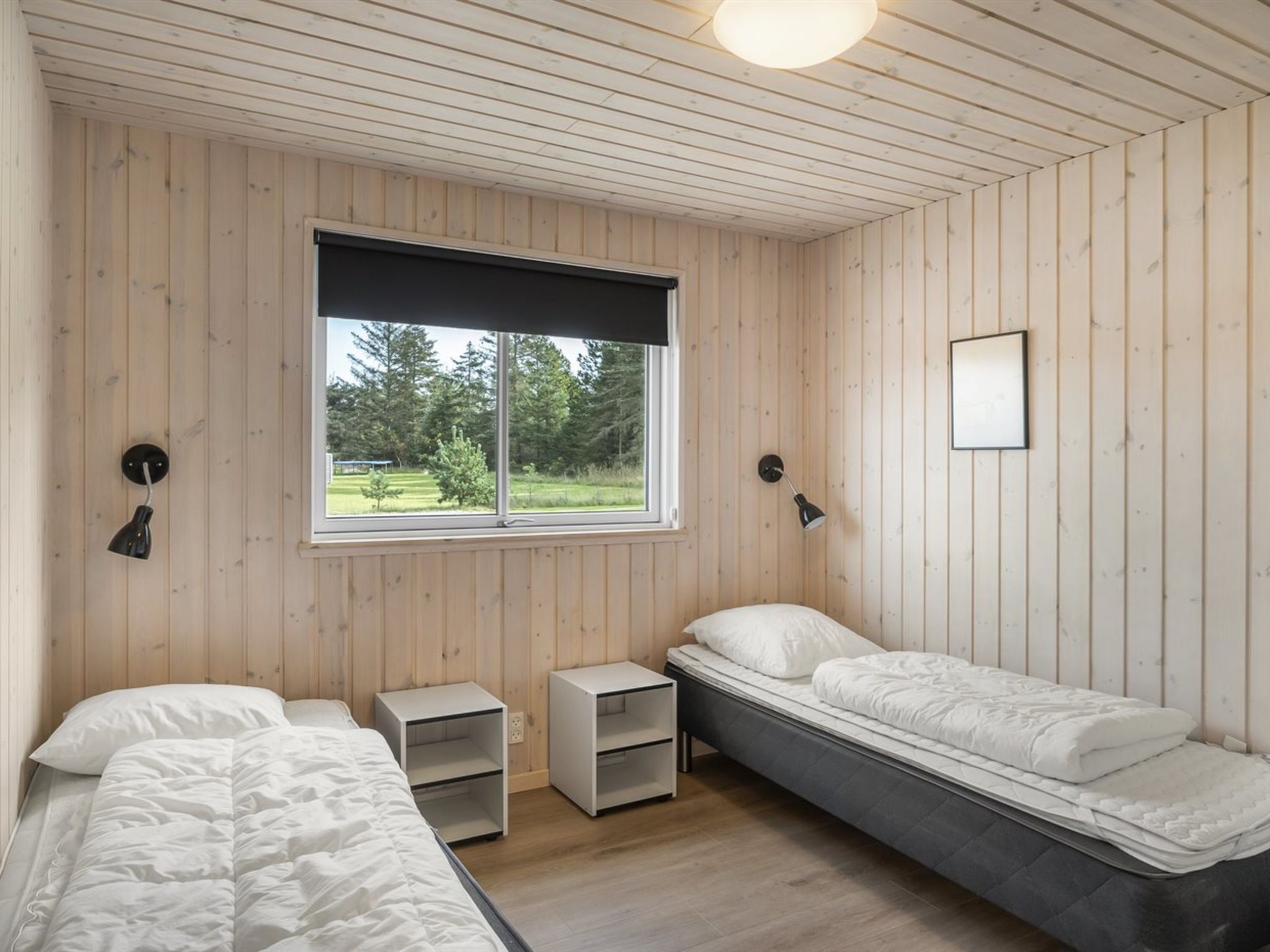 A room with two beds, a window with a view of greenery, and a ceiling light.