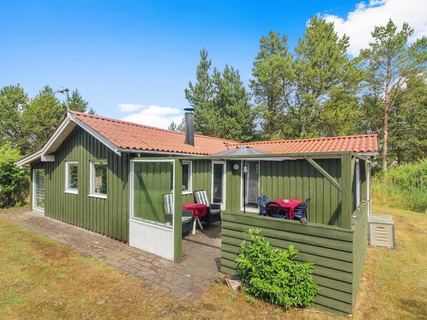Une petite cabane en bois d’un seul étage, avec un toit recouvert de tuiles rouges, une véranda et un petit abri attenant.