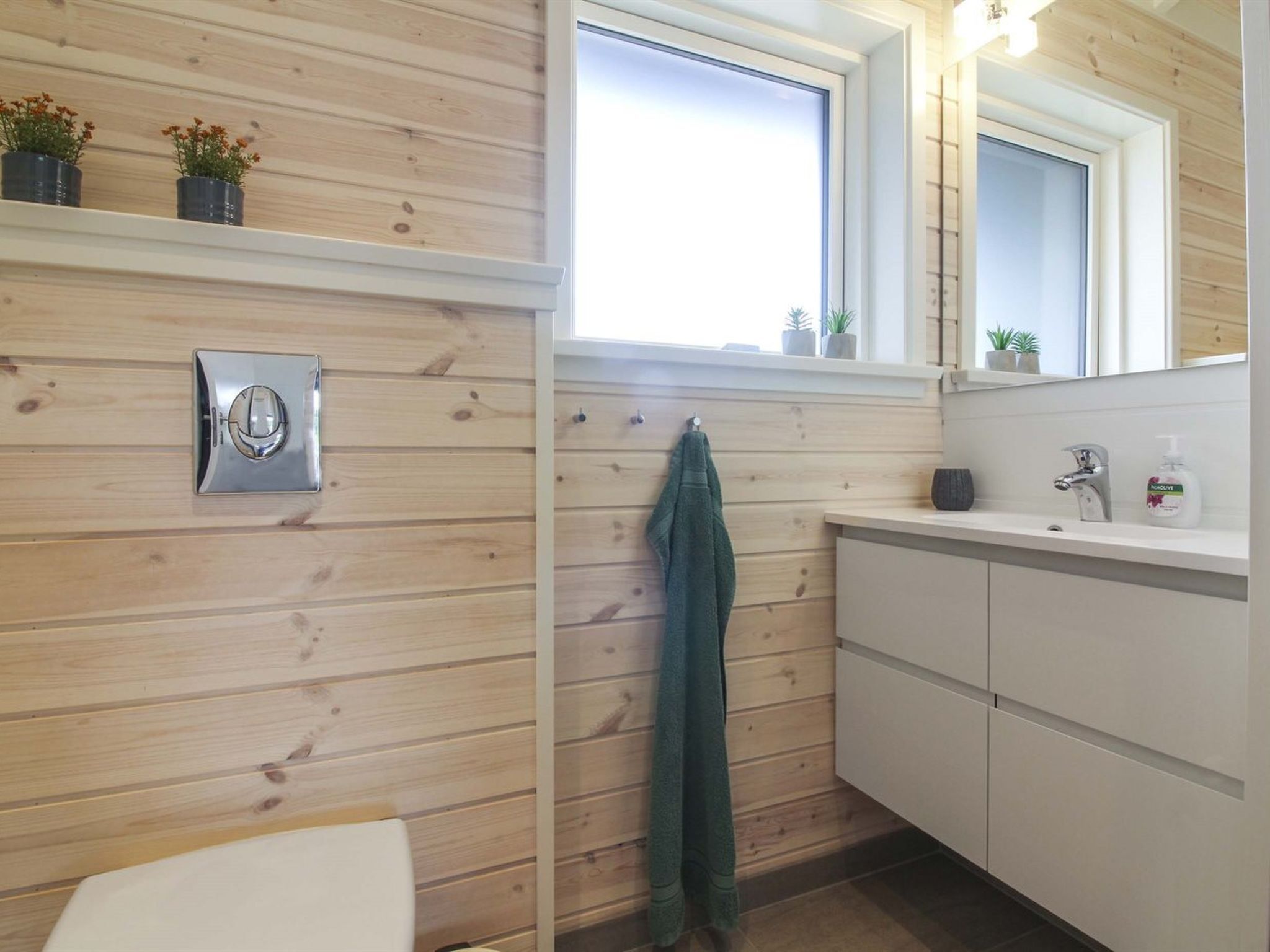 Ein Badezimmer mit Holzwandverkleidung, einem Fenster, einem an der Wand hängenden Handtuch, einem Waschbecken mit Spiegel und einer Toilette.