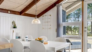 Un coin repas avec une table et des chaises blanches, un luminaire suspendu et une vue à travers une porte vitrée.