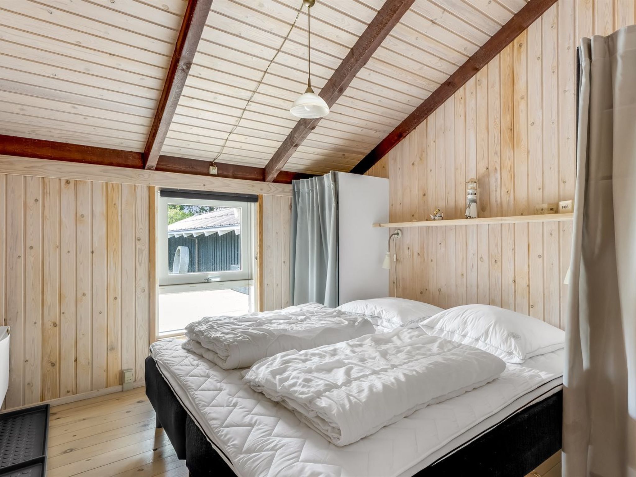 Une chambre avec un plafond en bois, un lit avec une literie blanche, une fenêtre avec des rideaux et une étagère ornée d’objets décoratifs.